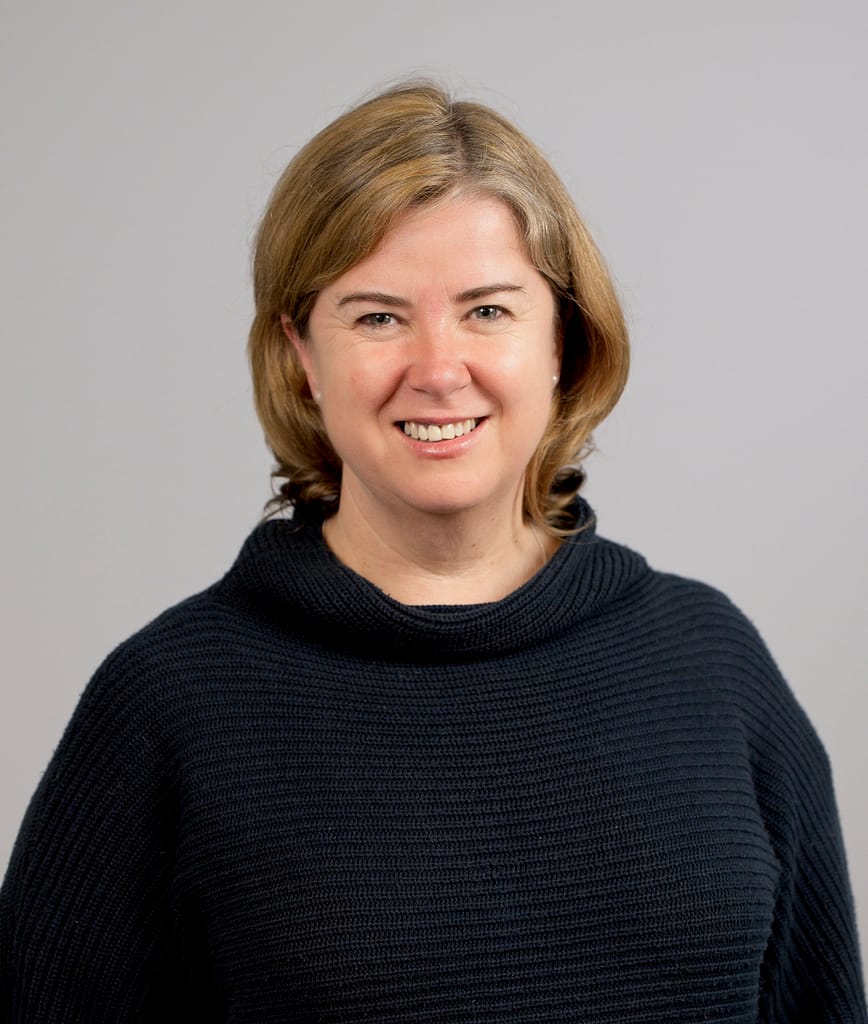 Heashot of Julie Arblaster. Julie is smiling at the camera and wearing a dark blue turtleneck jumper.